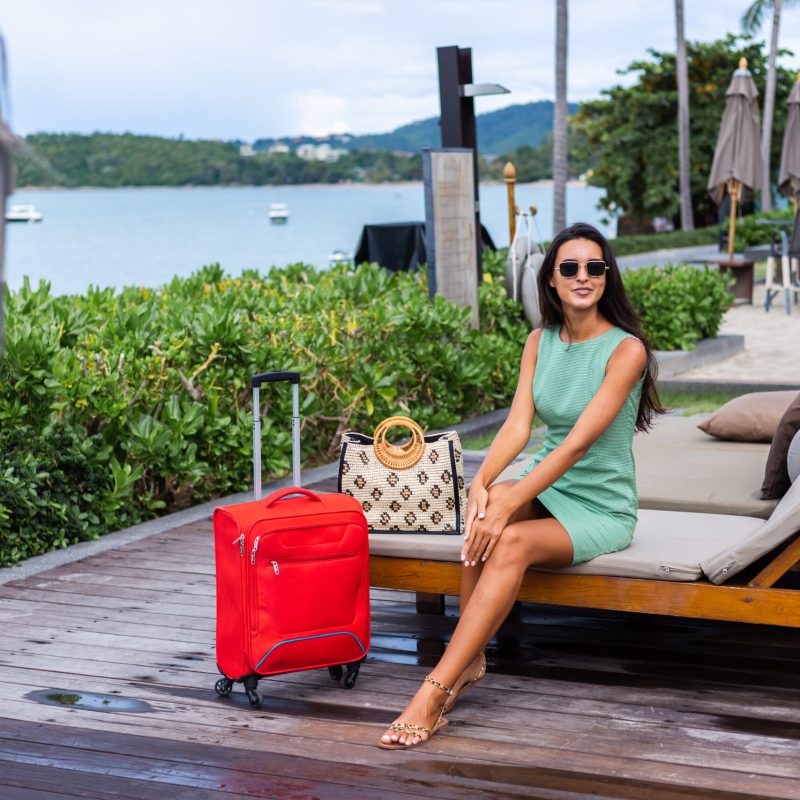 mulher-caucasiana-feliz-de-cabelos-longos-e-elegantes-turistas-em-um-vestido-com-mala-vermelha-fora-hotel